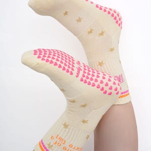 May include: A pair of cream-colored socks with pink heart-shaped grips on the soles and heels. Gold star patterns are scattered across the socks. The cuff features the text "The Life of a Barre Girl" with orange and pink stripes.