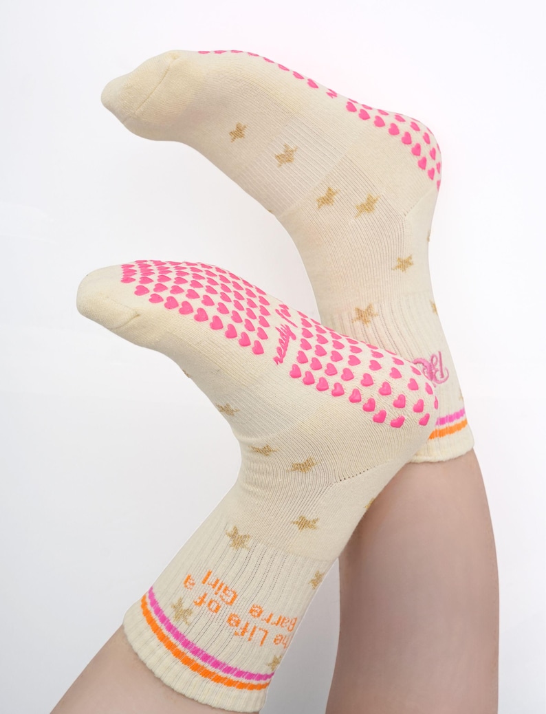 May include: A pair of cream-colored socks with pink heart-shaped grips on the soles and heels. Gold star patterns are scattered across the socks. The cuff features the text "The Life of a Barre Girl" with orange and pink stripes.