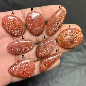 Puede incluir: Una colecci&oacute;n de ocho piedras preciosas de piedra solar pulidas en varias formas ovaladas y de l&aacute;grima. Las piedras son de un color rojo anaranjado c&aacute;lido con inclusiones brillantes, sostenidas en una mano.