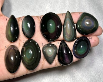 Op de afbeelding: Tien gepolijste regenboog-obsidiaan cabochons, genummerd van één tot tien, met concentrische banden van iriserend groen, paars en zwart. De stenen hebben ovale, druppelvormige en afgeronde driehoekige vormen met gladde, reflecterende oppervlakken.