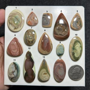 Puede incluir: Una colección de catorce piedras preciosas cabujón pulidas y multicolores dispuestas en un tablero blanco, numeradas del uno al catorce, con patrones en espiral en tonos crema, marrón, rosa y verde, junto a una moneda de cuarto de dólar.