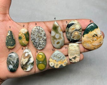 Puede incluir: Once cabujones de piedras preciosas pulidas en varias formas y colores, incluyendo verde, marrón, amarillo y gris. Las piedras se sostienen en una mano, mostrando sus patrones y texturas únicos. Ideal para la fabricación de joyas o coleccionar.