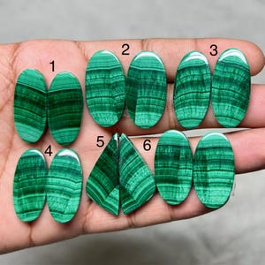 May include: A collection of six pairs of polished malachite gemstones, each displaying unique patterns of dark and light green bands. The stones are oval and triangular, showcasing natural mineral formations. The gemstones are displayed against a neutral background.