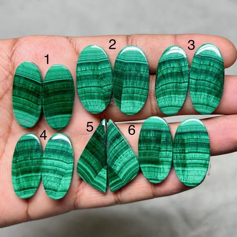 May include: A collection of six pairs of polished malachite gemstones, each displaying unique patterns of dark and light green bands. The stones are oval and triangular, showcasing natural mineral formations. The gemstones are displayed against a neutral background.