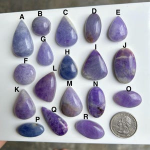 May include: A collection of polished gemstones in various shapes and shades of purple and blue. The gemstones include teardrop, oval, and round shapes. A US quarter is included for size reference.