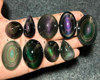 Op de afbeelding: Negen gepolijste regenboog-obsidiaan cabochons genummerd van één tot negen, met concentrische banden van iriserende paarse, groene en gouden kleuren op een zwarte basis. De stenen zijn glad, afgerond en ovaal of druppelvormig van vorm.