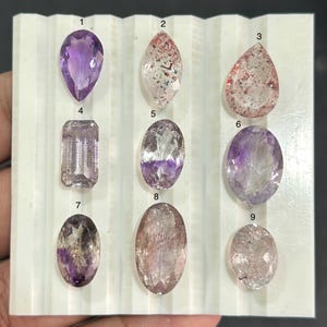 May include: A selection of nine polished gemstones in different shapes and colours, presented on a white surface. The gemstones include amethyst, quartz with red inclusions, and other stones with purple and clear hues. The shapes vary from teardrop to oval and rectangular.