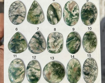 May include: Fifteen polished moss agate gemstones in various shapes, including oval, teardrop, and round. The stones display a mix of green, white, and brown patterns, with visible inclusions. Each stone is numbered for identification.