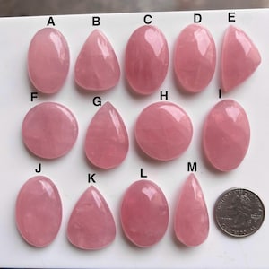 Puede incluir: Una colección de piedras preciosas de cuarzo rosa pulidas en varias formas, incluyendo ovaladas, de lágrima y redondas. Las piedras son de un color rosa suave y se muestran sobre una superficie blanca. Se incluye una moneda para referencia de tamaño.