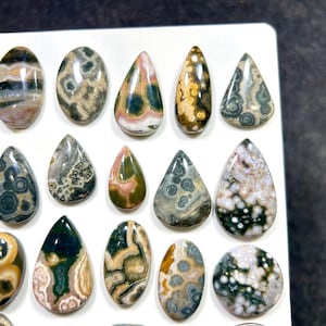 May include: A collection of polished, smooth gemstones in various shapes including teardrops and ovals. The stones feature intricate patterns in shades of green, brown, white, pink, and gray, with distinct circular and organic markings throughout each piece.