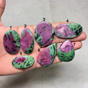 Pode incluir: Sete pedras preciosas polidas de rubi zoisite em várias formas, incluindo oval, gota e forma livre. As pedras apresentam uma mistura de tons rosa escuro e verde, com manchas pretas. As pedras preciosas são seguradas em uma mão.