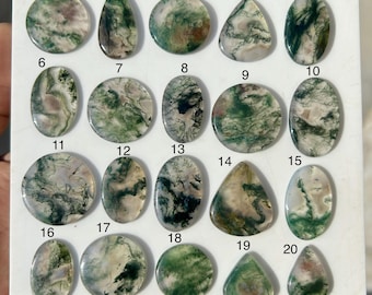 May include: A collection of twenty moss agate gemstones in various shapes, including oval, teardrop, and round. The stones display a mix of green, white, and brown patterns, with visible inclusions. The gemstones are arranged on a white surface.