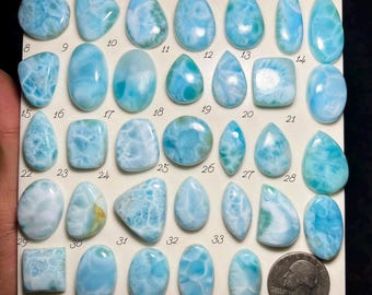 May include: A collection of thirty-three polished Larimar cabochons in various shapes including oval, teardrop, and square, arranged on a white display board with numbered labels from 1 to 33 and a US quarter coin for scale reference.