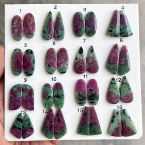 Puede incluir: Una colección de dieciséis pares de cabujones de rubí en zoisita pulidos dispuestos sobre un tablero blanco. Cada par presenta patrones distintos de inclusiones de rubí rojo magenta en zoisita verde con motas negras.