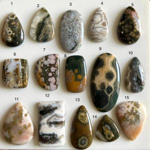 May include: Fifteen polished gemstone cabochons in various shapes, including teardrop, oval, and rectangular. The stones display a range of colors, such as green, brown, white, and gray, with unique patterns and textures. Ideal for jewelry making.