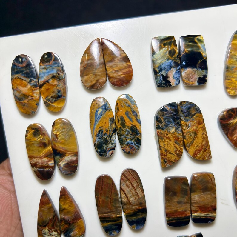 Op de afbeelding: Een verzameling gepolijste edelstenen in diverse vormen met wervelende patronen in diepblauw, goudgeel en bruin. Deze cabochons zijn in paren gerangschikt op een witte ondergrond en tonen gevarieerde ovale, druppelvormige en rechthoekige vormen.