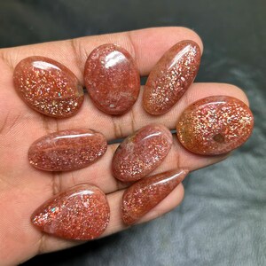 Puede incluir: Una colecci&oacute;n de ocho piedras preciosas de piedra solar pulidas en varias formas ovaladas y de l&aacute;grima. Las piedras son de un color rojo-naranja c&aacute;lido con inclusiones brillantes, sostenidas en una mano.