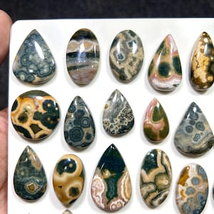 May include: A collection of polished, multi-colored cabochons in various shapes including teardrops and ovals. The stones feature intricate patterns of green, brown, cream, and pink, displaying natural orbicular and banded mineral inclusions.