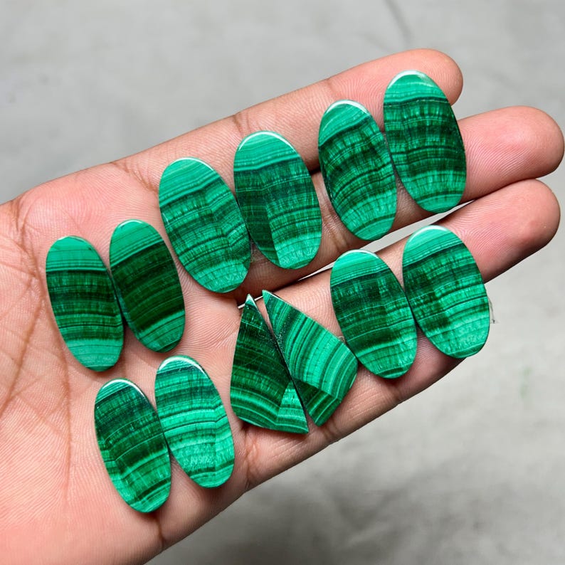 May include: A collection of polished malachite gemstones in various oval and triangular shapes. The stones display a vibrant green color with distinctive banded patterns, showcasing natural mineral formations. The gemstones are held in a hand.