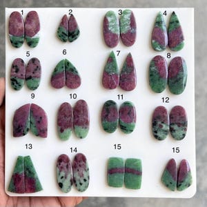May include: A collection of fifteen pairs of ruby zoisite cabochons in various shapes. The gemstones display a mix of deep red and green hues, with some having a marbled appearance. The shapes include ovals, teardrops, and triangles, suitable for jewelry making.