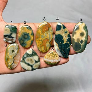 Può includere: Una collezione di otto pietre preziose lucide di varie forme. Le pietre presentano una gamma di colori, tra cui verde, marrone, giallo e crema, con motivi e texture unici. Le pietre preziose sono ovali, a goccia e rettangolari.