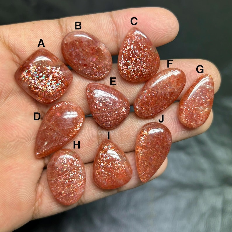 May include: A collection of ten sunstone gemstones in various shapes, including oval, teardrop, and diamond. The stones are a warm orange-red color with sparkling inclusions, held in a hand against a dark background. These gemstones are ideal for jewelry making.