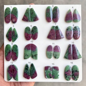 Puede incluir: Dieciséis pares de cabujones de rubí en zoisita pulidos dispuestos sobre un tablero blanco, numerados del uno al dieciséis. Cada par presenta patrones naturales distintos de inclusiones de rubí rojo magenta vibrante en piedra zoisita verde.