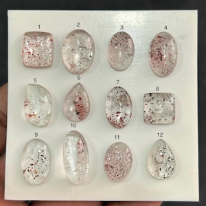 Peut inclure: Une collection de douze pierres précieuses en quartz clair avec des inclusions de rutile rouge, chacune taillée dans des formes différentes. Les pierres sont exposées sur une surface blanche. Les formes comprennent ovale, goutte et carré.