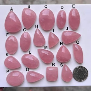 Könnte beinhalten: Eine Sammlung polierter, rosa Rosenquarz-Edelsteine in verschiedenen Formen, darunter oval, tropfenförmig und unregelmäßig. Eine Münze dient als Größenvergleich. Die Edelsteine sind auf einer weißen Oberfläche angeordnet.