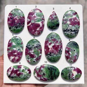 May include: Eleven polished ruby zoisite gemstones are displayed. The stones are in various shapes, including oval and teardrop, and feature a mix of green, deep red, and white colours. The gemstones are numbered 1 to 11.