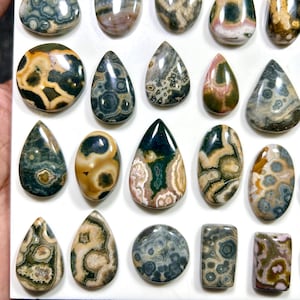 May include: A collection of polished ocean jasper cabochons in various shapes including teardrops, ovals, and rectangles. The stones feature intricate orbicular patterns in shades of green, brown, cream, pink, and grey with a smooth, glossy finish.