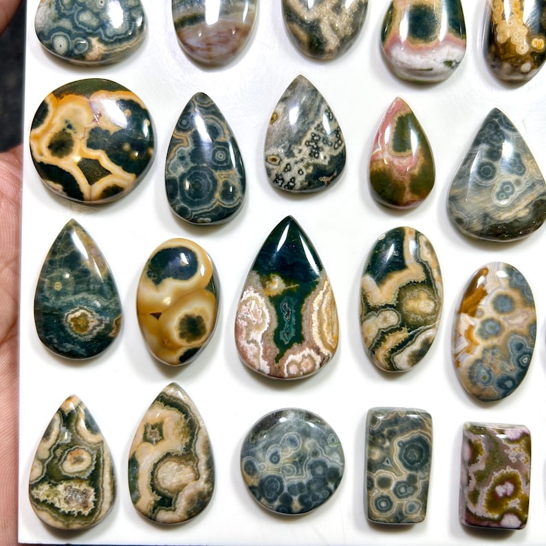 May include: A collection of polished ocean jasper cabochons in various shapes including teardrops, ovals, and rectangles. The stones feature intricate orbicular patterns in shades of green, brown, cream, pink, and grey with a smooth, glossy finish.