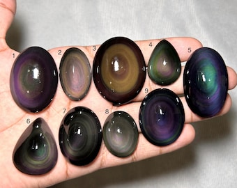 Op de afbeelding: Negen gepolijste regenboog-obsidiaan cabochons in verschillende ovale en druppelvormen, elk met concentrische banden van iriserende paarse, groene en gouden kleuren. De stenen zijn genummerd van één tot negen en hebben een glad, reflecterend oppervlak.