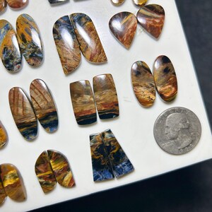 Op de afbeelding: Een verzameling gepolijste, veelkleurige pietersiet edelstenen in diverse vormen zoals ovalen, rechthoeken en druppels, met wervelende patronen van blauw, goud en bruin, weergegeven naast een Amerikaanse kwartje voor de schaal.