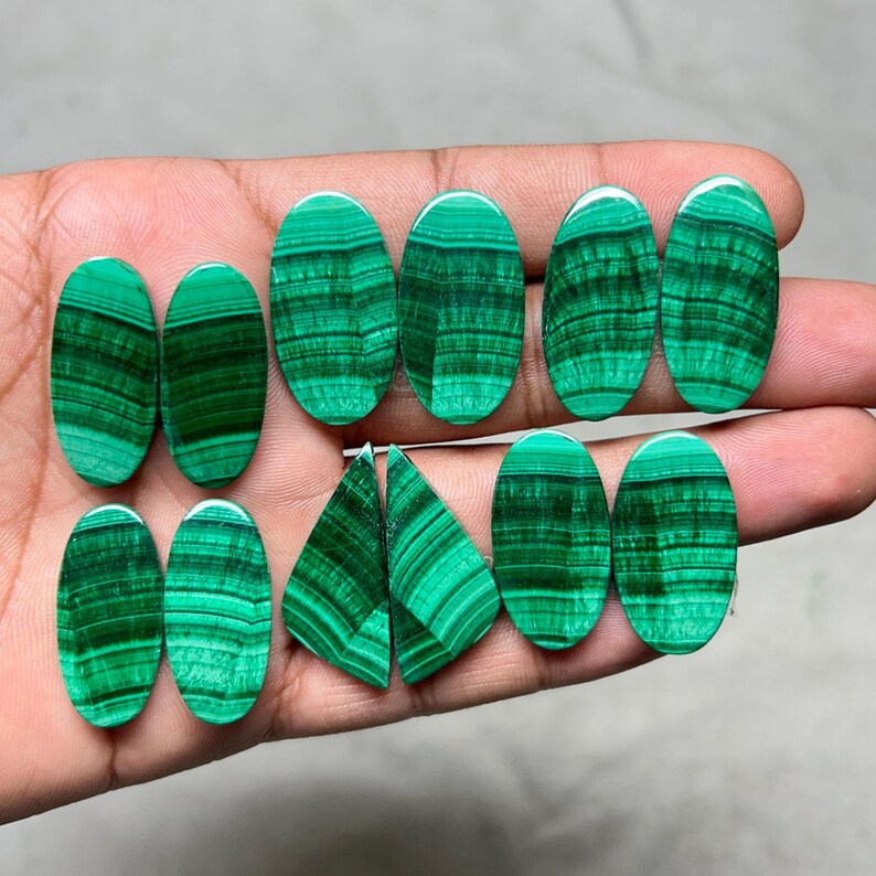 May include: A collection of polished malachite gemstones in various oval and triangular shapes. The stones display a vibrant green color with dark green banded patterns. The gemstones are held in a hand, showcasing their natural beauty and unique designs.