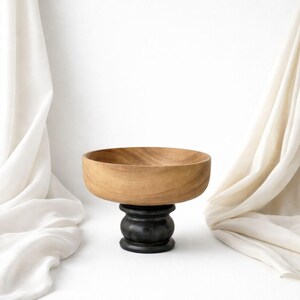 Puede incluir: Un cuenco de madera con acabado marrón claro se asienta sobre una base negra con forma de pedestal. El cuenco tiene un diseño ancho y poco profundo. El fondo es blanco con tela drapeada a ambos lados.