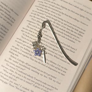Pu&ograve; includere: Segnalibro in metallo argentato con un ciondolo decorativo. Il ciondolo include una corona, una spada e un fiore viola. Il segnalibro &egrave; inserito in un libro.