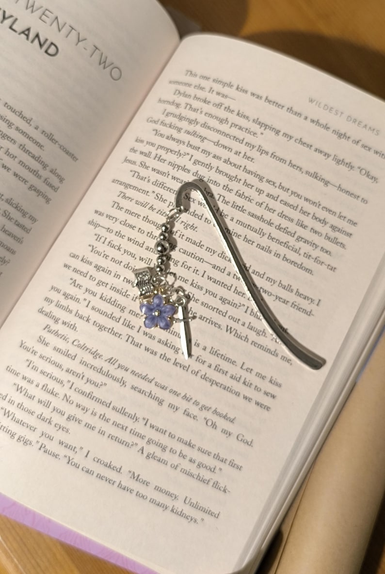 Pu&ograve; includere: Segnalibro in metallo argentato con un ciondolo decorativo. Il ciondolo include una corona, una spada e un fiore viola. Il segnalibro &egrave; inserito in un libro.
