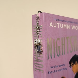 Pu&ograve; includere: Un segnalibro in metallo argentato con un ciondolo decorativo con un libro in miniatura, una chiave e un accento blu. Il segnalibro &egrave; attaccato a un libro viola intitolato "NIGHTSHADE" di Autumn Woods.