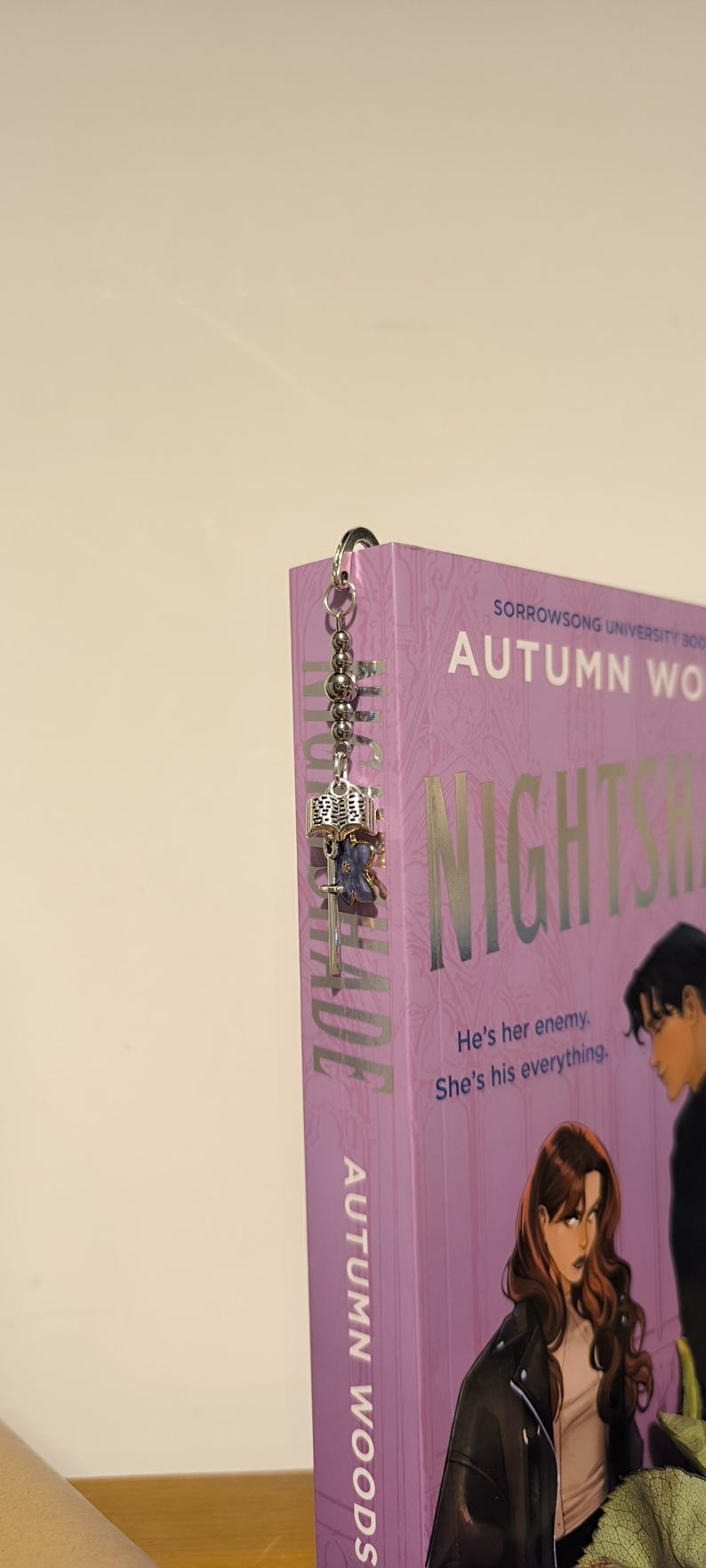 Pu&ograve; includere: Un segnalibro in metallo argentato con un ciondolo decorativo con un libro in miniatura, una chiave e un accento blu. Il segnalibro &egrave; attaccato a un libro viola intitolato "NIGHTSHADE" di Autumn Woods.
