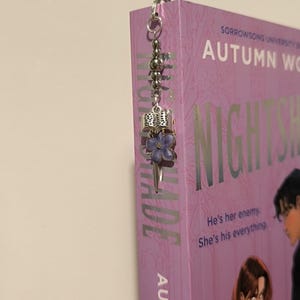 Pu&ograve; includere: Un segnalibro argentato con un ciondolo a forma di fiore viola e dettagli di perline &egrave; attaccato a un libro. Il libro ha una copertina viola con il titolo "NIGHTSHADE" e il nome dell'autore "AUTUMN WO". &Egrave; visibile anche il testo "He's her enemy. She's his everything."