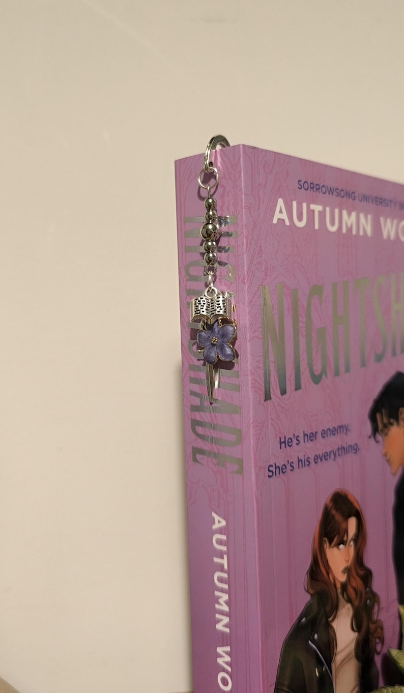 Pu&ograve; includere: Un segnalibro argentato con un ciondolo a forma di fiore viola e dettagli di perline &egrave; attaccato a un libro. Il libro ha una copertina viola con il titolo "NIGHTSHADE" e il nome dell'autore "AUTUMN WO". &Egrave; visibile anche il testo "He's her enemy. She's his everything."