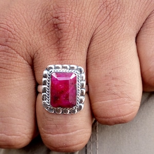 May include: A silver ring featuring a large, rectangular, deep red gemstone. The gemstone is set within a decorative, textured silver frame. The ring is shown on a person's finger.