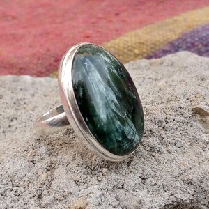 May include: A silver ring featuring a large, oval-shaped gemstone with a dark green, striated pattern. The gemstone is set in a polished silver bezel, creating a striking contrast. The ring is displayed on a textured, gray surface.