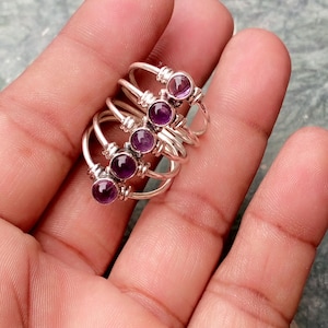 May include: A collection of silver rings, each with a round, deep purple gemstone. The rings are stacked together, highlighting their delicate design and the vibrant colour of the stones. The rings are set against a neutral background.
