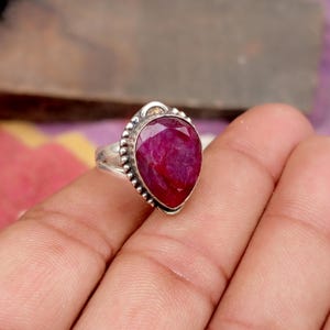 May include: A silver ring featuring a large, pear-shaped, faceted ruby gemstone. The ruby is a deep, vibrant red color. The ring has a detailed, beaded border around the gemstone and a simple band.