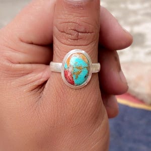May include: A silver ring featuring an oval gemstone with turquoise, red, and gold hues. The ring is displayed on a finger, highlighting the colourful stone. The band of the ring is plain silver.