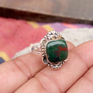 Könnte beinhalten: Ein Silberring mit einem quadratischen, dunkelgrünen Stein mit roten Einschlüssen. Der Ring hat eine verzierte, detaillierte Fassung mit floralen und perlenartigen Akzenten. Der Edelstein ist etwa 2 cm breit.