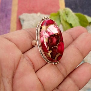 May include: A silver ring featuring a large, oval-shaped stone with a mosaic pattern of red, white, and gold hues. The stone is set in a silver band with a textured border.
