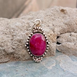 May include: A silver pendant featuring a large, oval-shaped, faceted ruby gemstone. The ruby is a vibrant shade of red. The pendant has a decorative silver border with small, round accents. The bail is a simple loop.